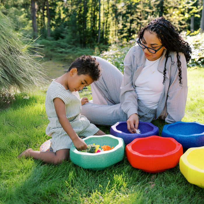 A woman and a young boy sit on grass outdoors, sorting colorful StepPebbles and a FREE Balance Board | Primary in large bowls. Sunlight filters through trees, creating a calm and playful indoor-outdoor scene.