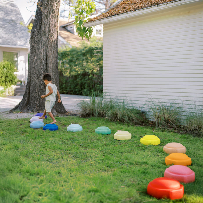 A young child enjoys indoor-outdoor play on grass, stepping on StepPebbles and a FREE Balance Board | Primary, arranged in a curved line beside a white house and tree.