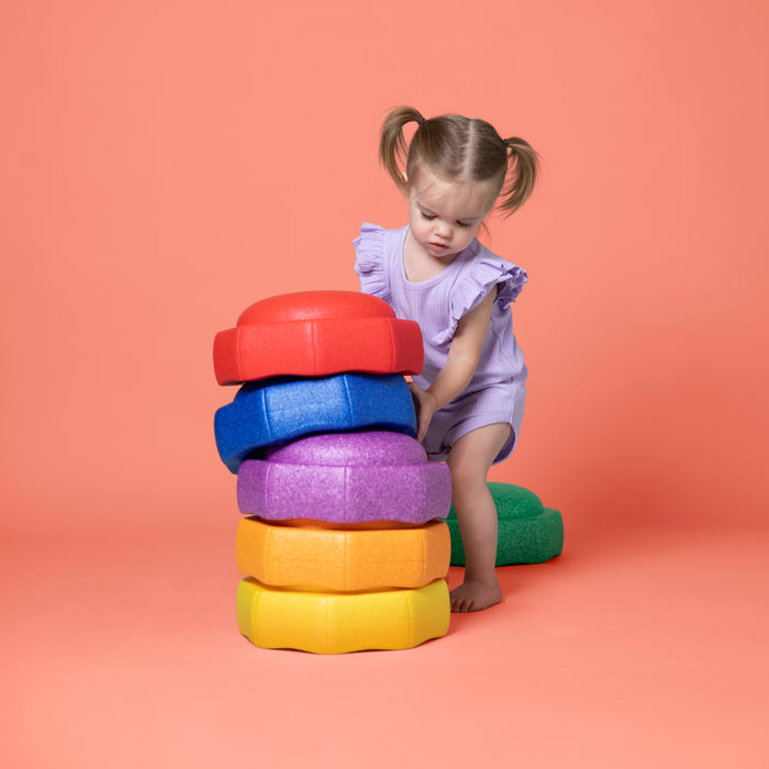 A young child in a light purple outfit stacks vibrant StepPebbles and a FREE Balance Board | Primary, ideal for kids' balance play indoors or outdoors, against a peach-colored background.