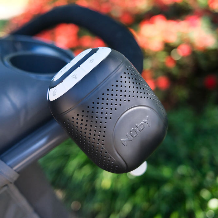 A Nuby Stroller Rocker in black is attached to a stroller handle outdoors, with soothing sounds playing amid blurred greenery and red flowers in the background.