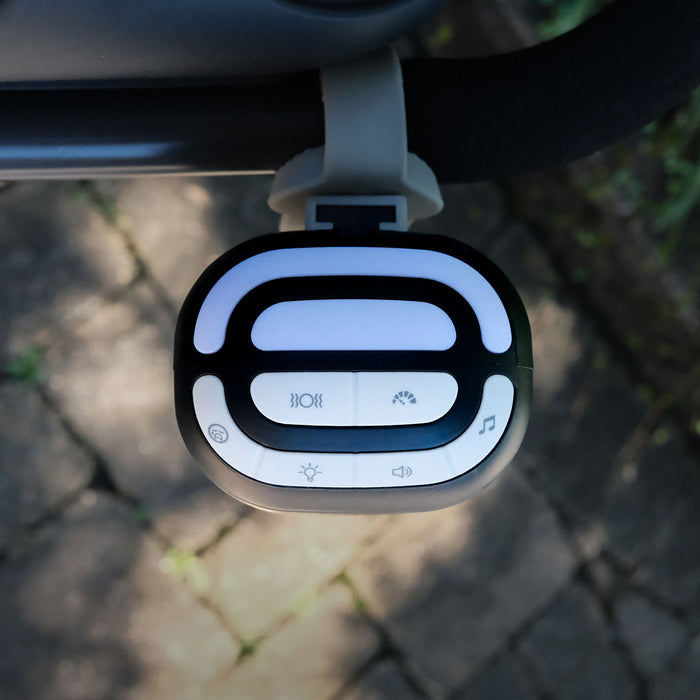 Close-up of Nuby Stroller Rocker's white and black control panel with four buttons—light, fan, soothing music, and brightness—mounted on the dark handlebar, set against a stone pavement background.