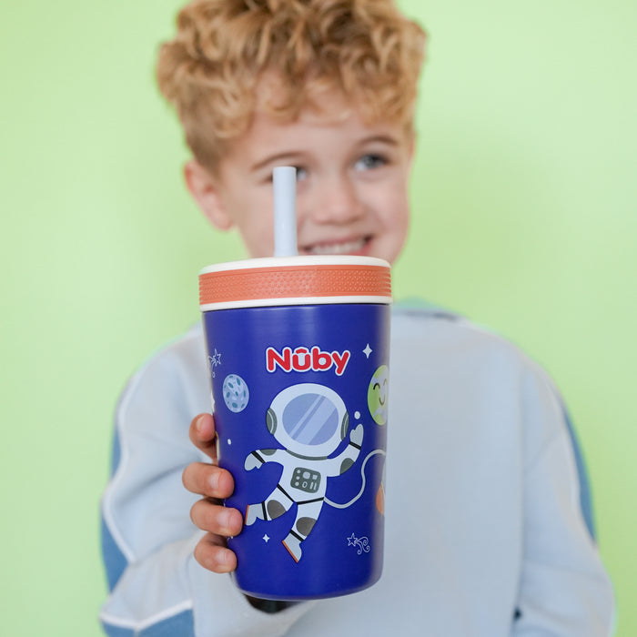 A smiling child with curly blond hair holds a Nuby Thirsty Kids Flexstraw Stainless Steel Cup | Astronaut, featuring an astronaut and planets design, standing against a light green background.