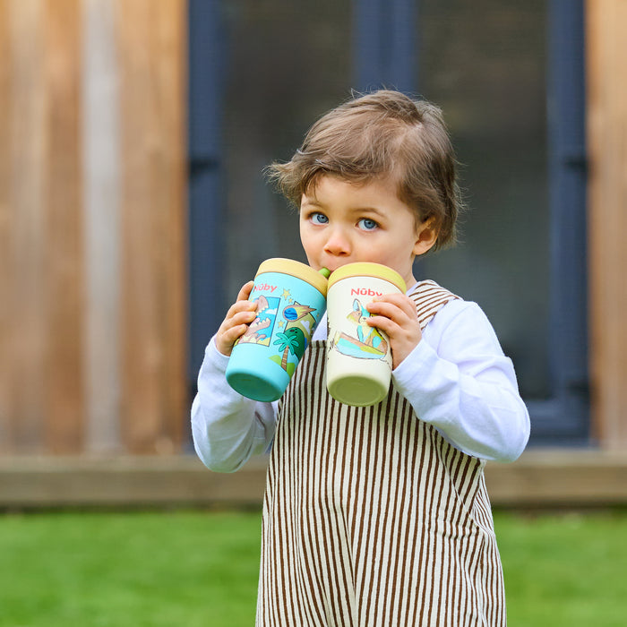 A young child with light brown hair in a striped pinafore and white shirt stands outside holding two Thirsty Kids Flexstraw Stainless Steel Cups | Dinosaurs, drinking from both. A blurred building and green grass are visible in the background.