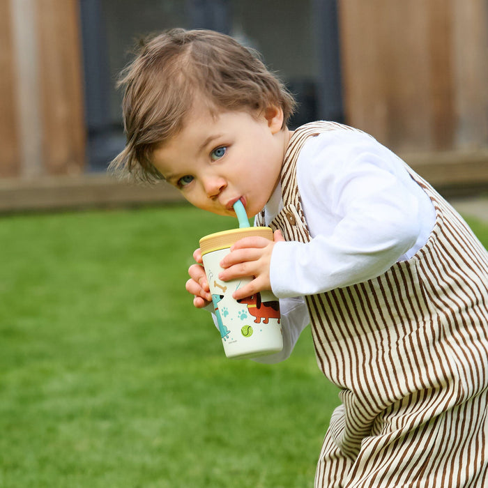 A young child with light brown hair, in a white shirt and striped overalls, stands on green grass outdoors, sipping from a Thirsty Kids Flexstraw Stainless Steel Cup | Dogs with a blue straw.
