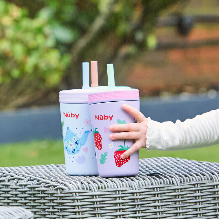 A child’s hand picks up the Thirsty Kids Flexstraw Stainless Steel Cup | Strawberries, next to a blue no-spill straw cup with whale designs, both resting on a wicker table outdoors.