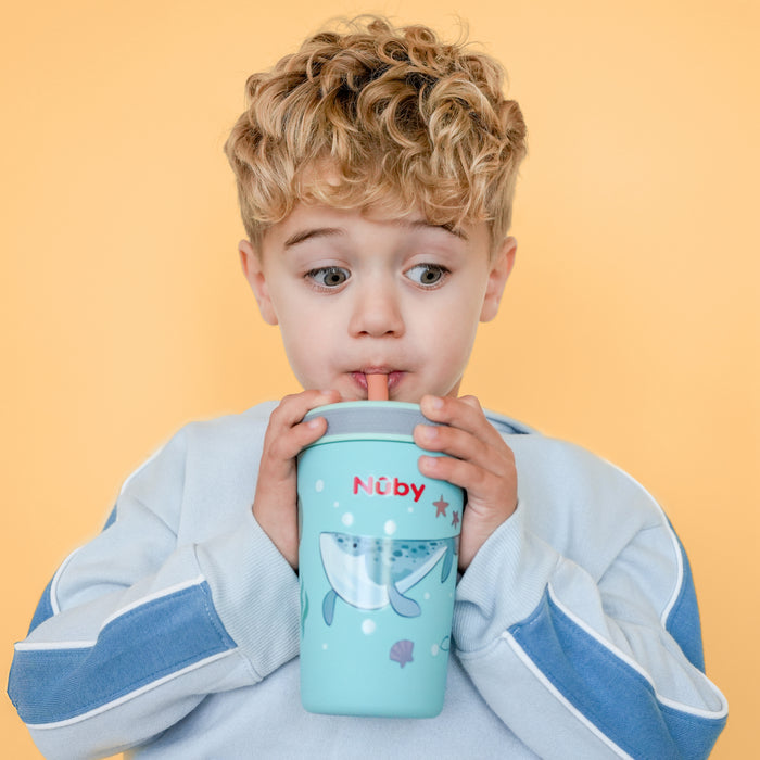 A young boy with curly blond hair and a surprised expression sips from a blue Nûby cup featuring a playful whale design, standing in front of a vibrant yellow background.