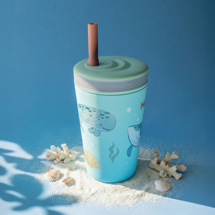 The Nuby Thirsty Kids Flexstraw Stainless Steel Cup | Whales, a light blue insulated kids cup with cartoon whales, a gray lid, and a brown straw, sits on sand with seashells and coral against a blue background with leafy shadows.