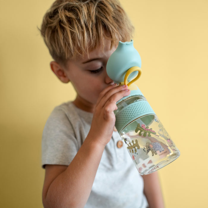 A young child with blond hair drinks from a Nuby US Thirsty Kids Flip-It BOLT Water Bottle | Mammoths, a clear kids cup with colorful mammoth illustrations and a silicone spout, set against a plain yellow background.