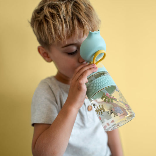 A young child with blond hair drinks from a Nuby US Thirsty Kids Flip-It BOLT Water Bottle | Mammoths, a clear kids cup with colorful mammoth illustrations and a silicone spout, set against a plain yellow background.