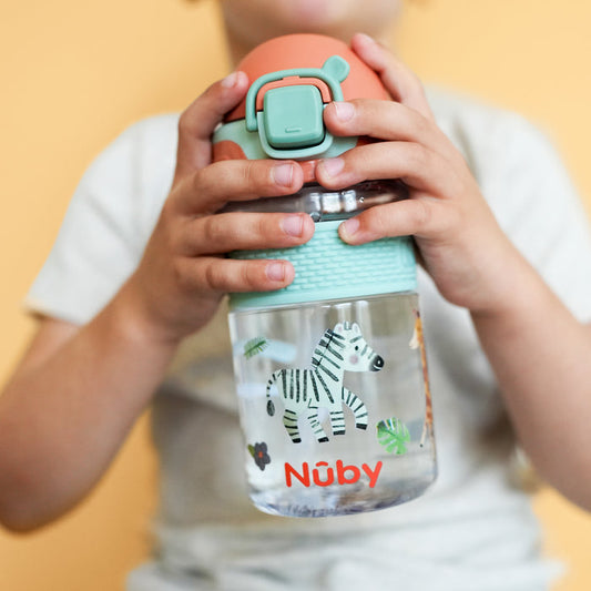 A young child holds a Nuby US Thirsty Kids Flip-It BOLT Water Bottle | Safari, featuring a green and orange lid with zebra design and silicone no-spill spout. The child wears a light shirt against a pale yellow background.