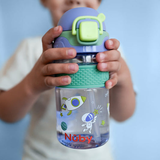 A child holds a clear Nuby US Thirsty Kids Flip-It BOLT Water Bottle | Space, decorated with an astronaut, rocket, planets, and stars. It has a green textured grip, a purple flip-top lid with latch, and a silicone spout for easy sipping.