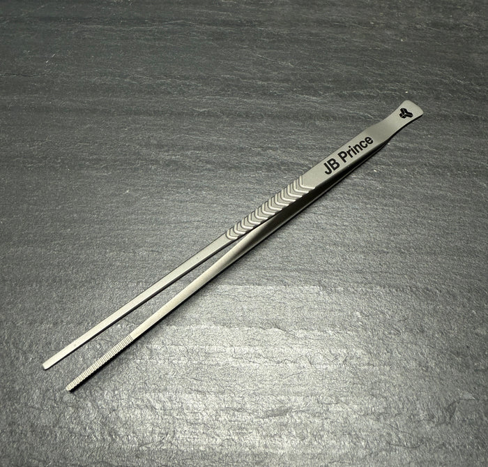 Metal titanium tweezers with 'JB Prince' engraving on a gray surface.