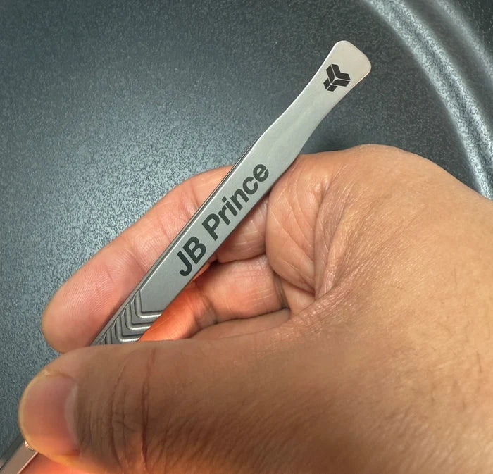 Hand holding a Titanium tweezer with 'JB Prince' branding against a gray background.