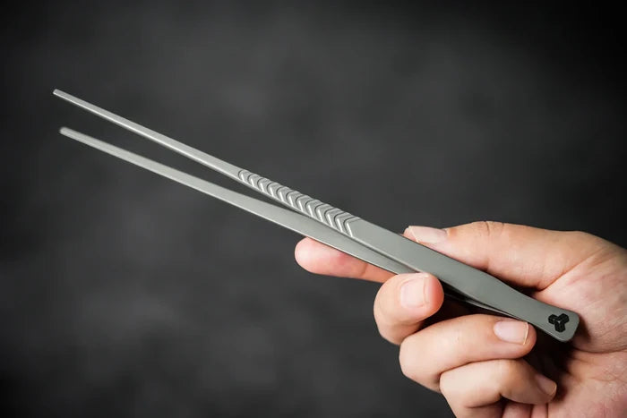 Hand holding a pair of Titanium Tweezers against a dark background.