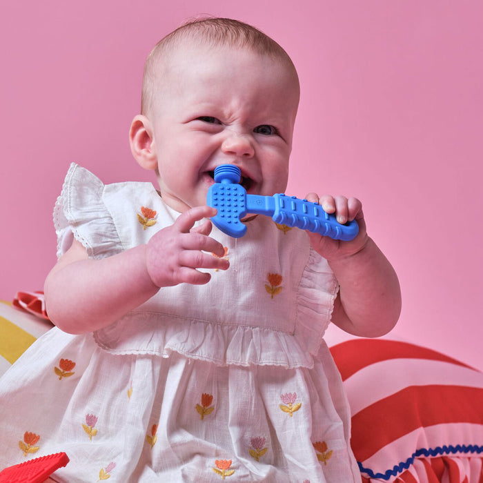 A baby in a white dress with flower patterns chews on a blue Tool Teether (4 Pack), sitting against a pink background with colorful cushions. These silicone teethers are perfect for soothing little gums.