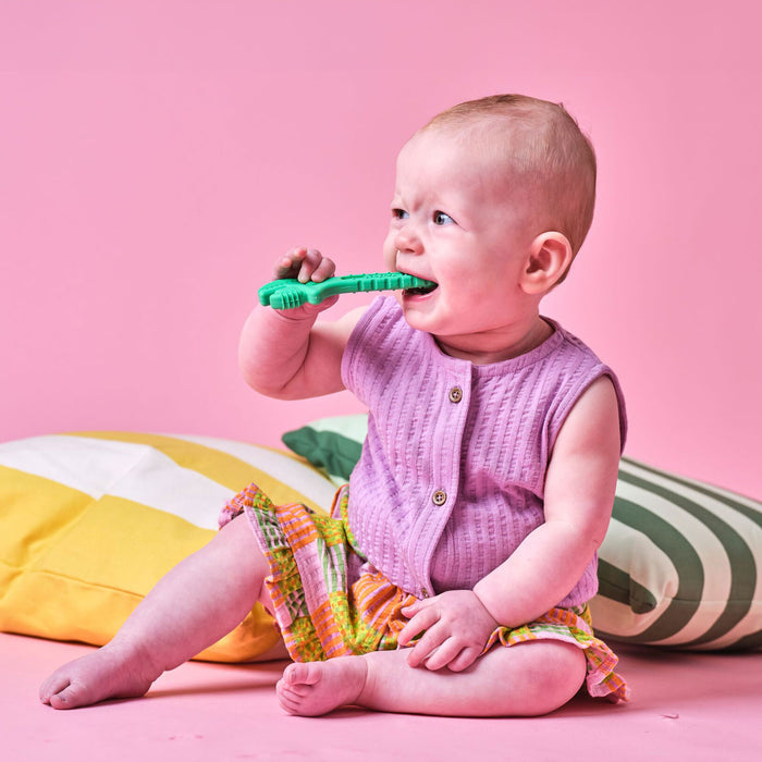 A baby in a purple top and colorful skirt sits on a pink floor, holding and chewing on a green Tool Teethers (4 Pack) silicone teether. Cushions with yellow and green stripes are behind the baby, set against a pink background.