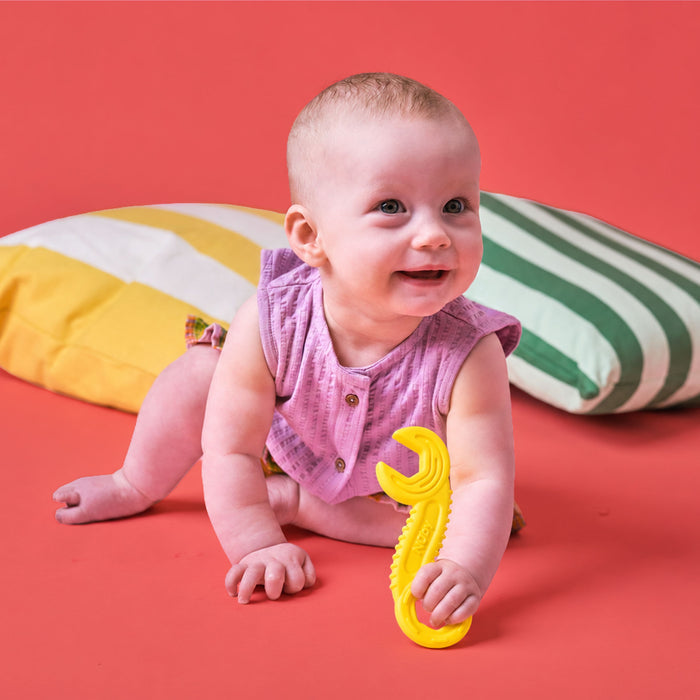 A smiling baby in a pink outfit sits on a red surface, holding a yellow Tool Teethers (4 Pack) teether shaped like a wrench. Two pillows—one with yellow and white stripes, the other green and white—are behind the baby.