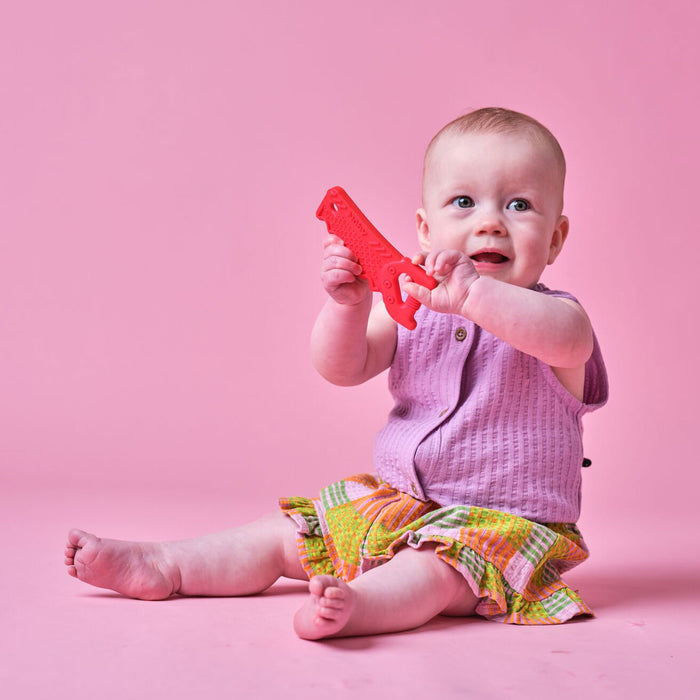 A smiling baby sits on a pink floor in a purple shirt and colorful shorts, holding a red Tool Teethers (4 Pack) silicone teether shaped like an alligator.
