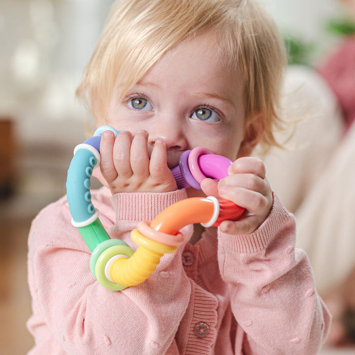 A blonde, blue-eyed toddler in a pink sweater holds and chews on the Twista Rattle Teether Toy | Colorful Rings for teething relief, gazing thoughtfully at their baby sensory toy.