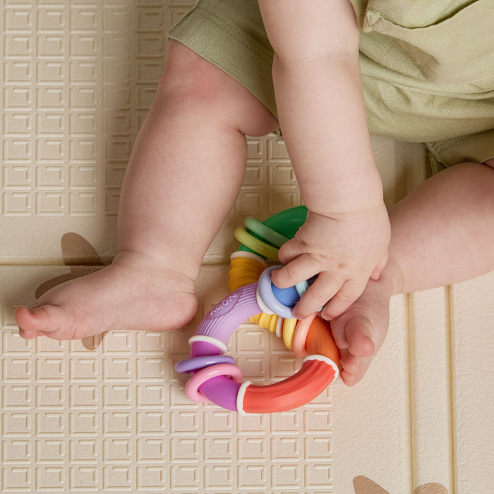 A baby in light green clothing sits on a textured mat with a simple square pattern, holding the Twista Rattle Teether Toy | Colorful Rings in both hands. Their legs are visible.