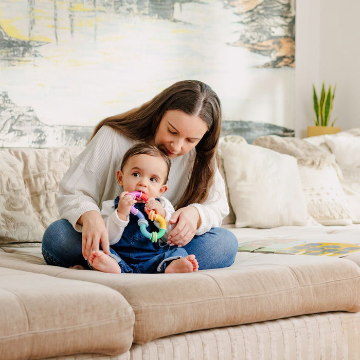 A woman sits on a beige couch with a baby in her lap. The baby holds the Twista Rattle Teether Toy | Colorful Rings, enjoying soothing teething relief and sensory play amid soft pillows and a plant for a cozy home vibe.