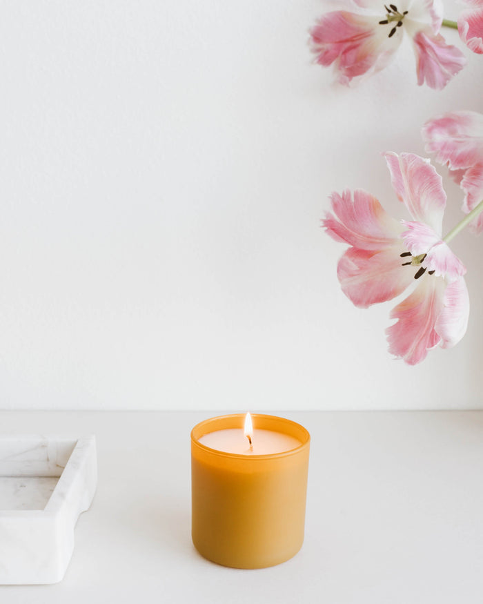 Lit amber glass candle on white surface near pink tulip flowers with white wall background, featuring Vetiver + Tonka Dignity Series Glass Jar Soy Candle | Calyan Wax Co. - 5.3 oz