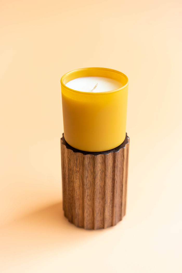 Vetiver + Tonka Dignity Series Glass Jar Soy Candle | Calyan Wax Co. - 5.3 oz displayed in a warm yellow glass jar atop a textured wooden stand against a soft beige background.
