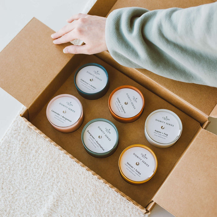 Open cardboard box containing six colored glass jar candles with white labels, including Vetiver + Tonka Dignity Series Glass Jar Soy Candle | Calyan Wax Co. - 5.3 oz, on a light surface with a hand reaching in.
