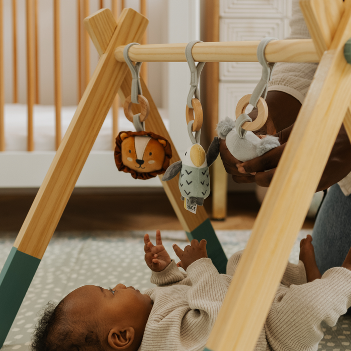 A baby lies on a play mat reaching up toward the Wooden Play Gym with hanging toys, while an adult adjusts one of the toys. A crib can be seen in the background.
