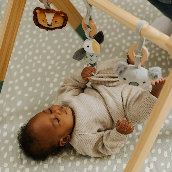 A baby in a beige outfit lies on a patterned mat, calmly reaching for plush animal toys hanging from a wooden activity gym, fully engaged with the colorful play space above.