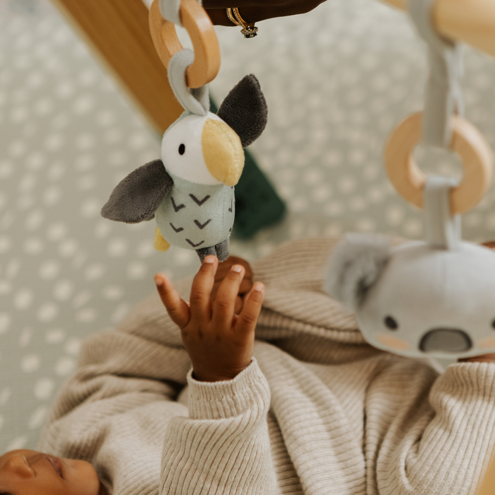 A baby reaches up toward hanging toys on a Wooden Play Gym, wearing a beige sweater. A plush koala sits nearby on a light dotted play mat.
